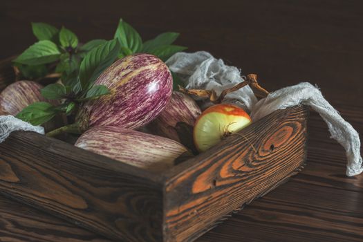 Purple graffiti eggplants, onion and green fresh basil in a wooden box in a vintage wooden background in rustic style, selective focus