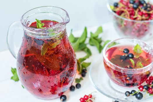 Red and black currant fruit drink in transparent glass cup. Clear glass vase with red and black currant berries on the white wooden background
