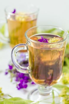 Linden herbal tea in a transparent grog glass with a linden blossom and bunch of herbs on the white wooden surface