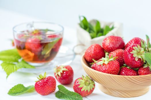 Fresh strawberry in wooden plate. Glass cup of summer tea with fresh strawberry. Green leaves. Fresh mint. White wooden table. Shallow depth of field.