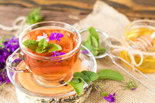 Glass cup of summer herbal tea with fresh mint and field larkspur. Jar of honey. Wooden table. Shallow depth of field.