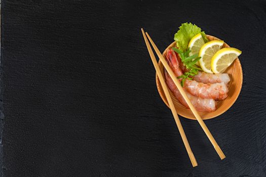 Cooked peeled shrimps with lemon slices, green lettuce and arugula leaves on a wooden plate with wooden chopsticks on a side of a plate. The plate is at the black stone countertop