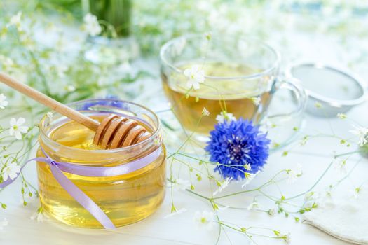 Honey in glass jars and cup of tea with white spring flowers and cornflower on windowsill. Shallow depth of field.