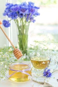 Honey in glass jars and cup of tea with white spring flowers and cornflower on windowsill. Shallow depth of field.