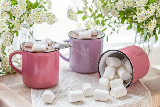 Marshmallows on top of hot cocoa in pink cups on the white napkin on the windowsill. Fresh white spring flowers are framing the image. Cozy home concept. Shallow depth of field