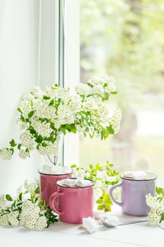 Hot cocoa with marshmallows in pink cups and fresh spring white flowers on the windowsill. Cozy home concept. Shallow depth of field.