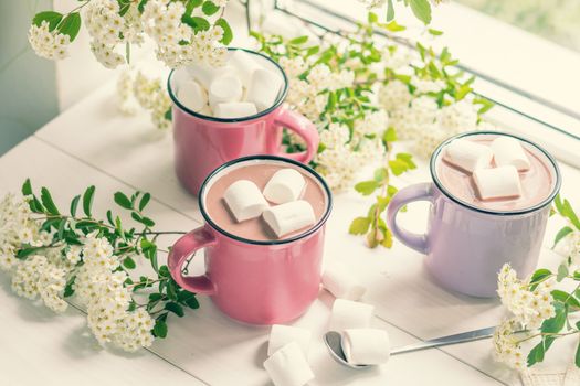 Hot cocoa with marshmallows in pink cups and fresh spring white flowers on the windowsill. Cozy home concept. Shallow depth of field.