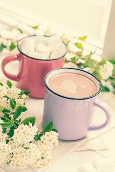 Hot cocoa with marshmallows in pink cups and fresh spring white flowers on the windowsill. Cozy home concept. Shallow depth of field.
