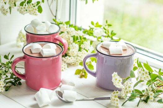 Hot cocoa with marshmallows in pink cups and fresh spring white flowers on the windowsill. Cozy home concept. Shallow depth of field.