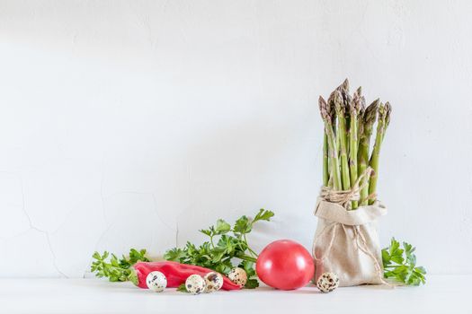 Bunches of fresh asparagus in a little sack, vegetables and quail eggs on the white cracked wall background. Copy space