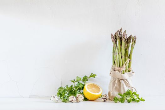 Bunches of fresh asparagus in a little sack, lemon and quail eggs on the white cracked wall background. Copy space