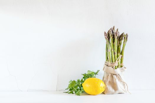 Bunches of fresh asparagus in a little sack, lemon and fresh parsley on the white cracked wall background. Copy space