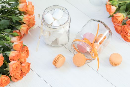 Orange mango or citrous macaroons and marshmallow in jars. Fresh little roses. Light wooden background. Sunlight. Coloring and processing photo with light vintage style. Shallow depth of field.