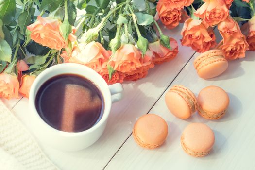 Orange mango or citrous macaroons, cup of coffee and fresh little roses on light wooden background. Sunlight. Coloring and processing photo with light vintage style. Shallow depth of field.