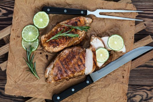 Sliced grilled chicken filet with herbs  on a paper on wooden background. Rosemary and sliced lime on a dark groundwork.