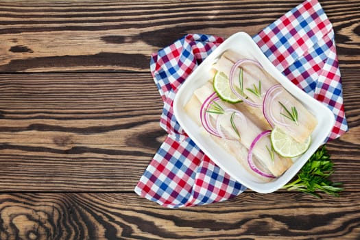 Sliced herring fillets, cut onion and lime on white plate. Checkered napkin. On wooden table.