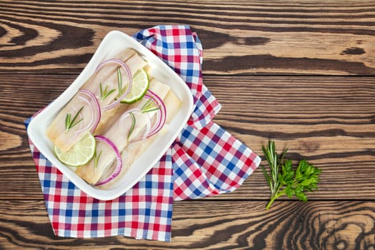Sliced herring fillets, cut onion and lime on white plate. Checkered napkin. On wooden table.