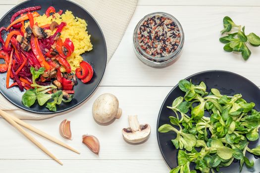 Healthy vegetarian diet concept. Rice and steamed vegetables, lamb's lettuce feldsalat on a black plates, chopsticks, napkin, garlic, mushrooms, raw multicolor rice in glass jar. White wooden table.