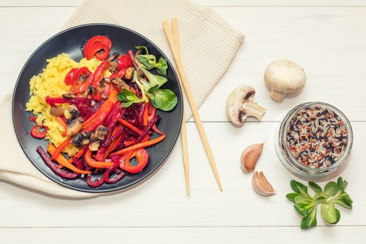 Healthy vegetarian diet concept. Rice and steamed vegetables, lamb's lettuce feldsalat on a black plate, chopsticks, napkin, garlic, mushrooms, raw multicolor rice in glass jar. White wooden table.