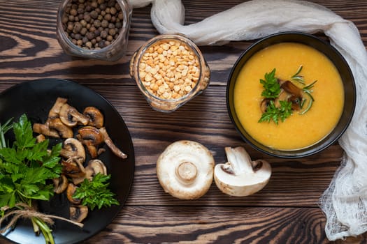 Pea soup with mushrooms in black plate. Fried mushrooms. Dried pea, pepper in glass jars.  Fresh parsley and rosemary. Dark wooden table. Toned.