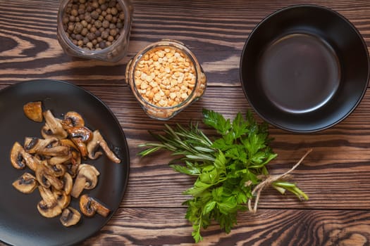 Fried mushrooms in black plate. Dried pea, pepper in glass jars.  Fresh parsley and rosemary. Dark wooden table. Toned.