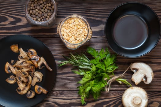 Fried mushrooms in black plate. Dried pea, pepper in glass jars.  Fresh parsley and rosemary. Dark wooden table. Toned.