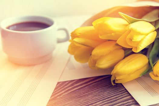 Beautiful spring music background. Cup of coffee, yellow tulips, musical page on a dark wooden background. Shallow depth of field. Coloring photo with soft focus in instagram style