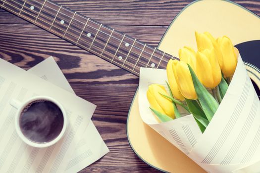 Beautiful spring background music. Guitar, yellow tulips, cup of coffee, musical page on a dark wooden background. Shallow depth of field. Coloring photo with soft focus in instagram style.