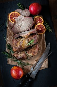 Sliced baked pork with herbs on a stone cutting board on wooden background.Grilled steak on a paper. Rosemary and sliced sicilian blood oranges  on a dark groundwork.