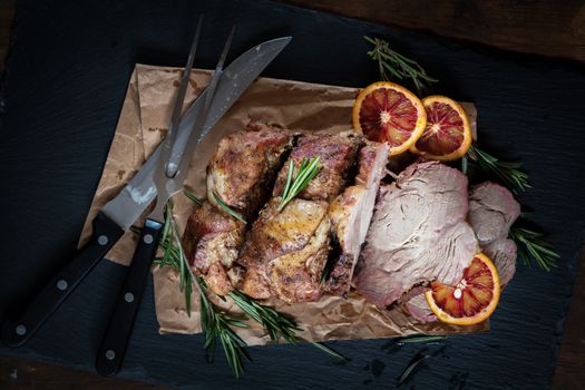 Sliced baked pork with herbs on a stone cutting board on wooden background.Grilled steak on a paper. Rosemary and sliced sicilian blood oranges  on a dark groundwork.