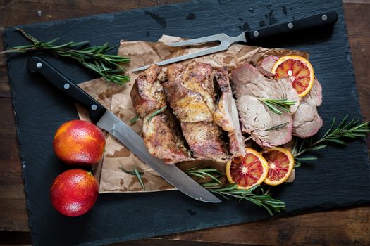 Sliced baked pork with herbs on a stone cutting board on wooden background.Grilled steak on a paper. Rosemary and sliced sicilian blood oranges  on a dark groundwork.