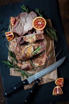 Sliced baked pork with herbs on a stone cutting board on wooden background.Grilled steak on a paper. Rosemary and sliced sicilian blood oranges  on a dark groundwork.
