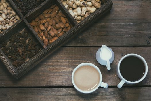 Cup of black coffee, cup of coffee with milk, cream. Almond, pistachios, anise, peanut and coffee beans in box. Dark wooden background. Beautiful vintage coffee groundwork. Coloring and processing photo.