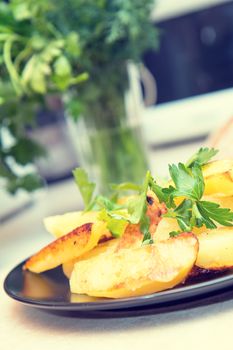 Delicious baked fried potatoes with parsley in black plate on table in the kitchen. Shallow depth of field. Coloring and processing photo.