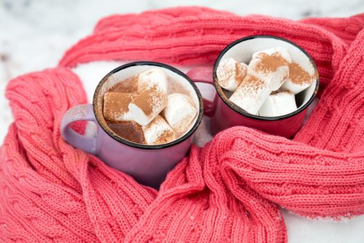 Hot chocolate with marshmallow in pink and violet two cups wrapped in a cozy winter pink scarf on the snow-covered table in the garden. Coloring and processing photo small depth of field