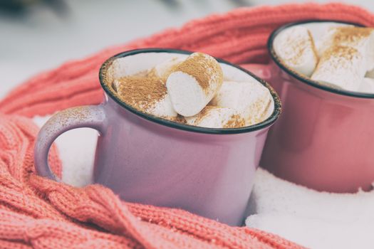 Hot chocolate with marshmallow in pink and violet two cups wrapped in a cozy winter pink scarf on the snow-covered table in the garden. Coloring and processing photo small depth of field