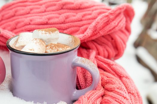 Hot chocolate with marshmallow in violet cup wrapped in a cozy winter pink scarf on the snow-covered table in the garden. Coloring and processing photo, selective focus, small depth of field