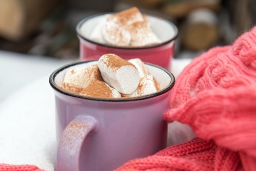 Hot chocolate with marshmallow in pink and violet two cups wrapped in a cozy winter pink scarf on the snow-covered table in the garden. Coloring and processing photo small depth of field