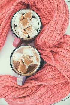Hot chocolate with marshmallow in pink and violet two cups wrapped in a cozy winter pink scarf on the snow-covered table in the garden. Coloring and processing photo, small depth of field