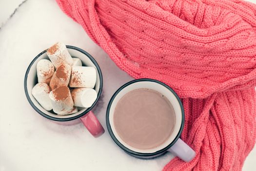 Hot chocolate with marshmallow in pink and violet two cups wrapped in a cozy winter pink scarf on the snow-covered table in the garden. Coloring and processing photo, small depth of field