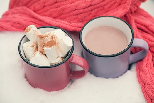 Hot chocolate with marshmallow in pink and violet two cups wrapped in a cozy winter pink scarf on the snow-covered table in the garden. Coloring and processing photo small depth of field