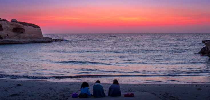 Three women waiting for the sunrise in front of the sea (Puglia region, South of Italy). Concept of frienship, travel and adventure.