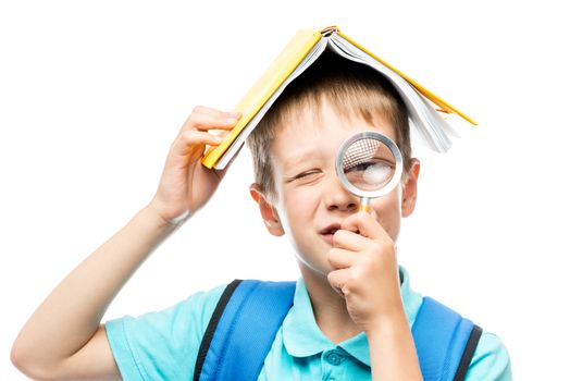 funny schoolboy with book on head looking through magnifier on white background