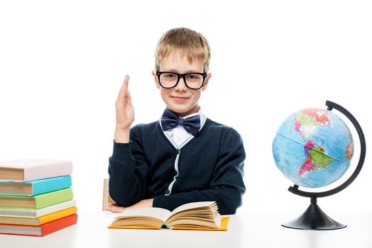 a schoolboy with glasses at a table with books and a globe pulls his hand to answer