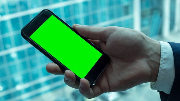 Businessman is holding black smartphone with green screen near the window in office