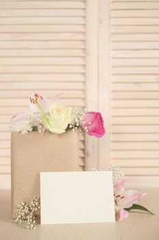 Flowers in paper bag and blank envelope on wooden background