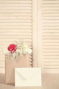 Flowers in paper bag and greeting envelope on wooden background