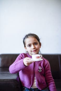 little girl plays, drinking tea on the sofa at home, lit by the window