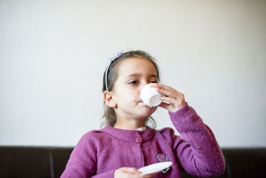 little girl plays, drinking tea on the sofa at home, lit by the window