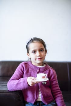 little girl plays, drinking tea on the sofa at home, lit by the window